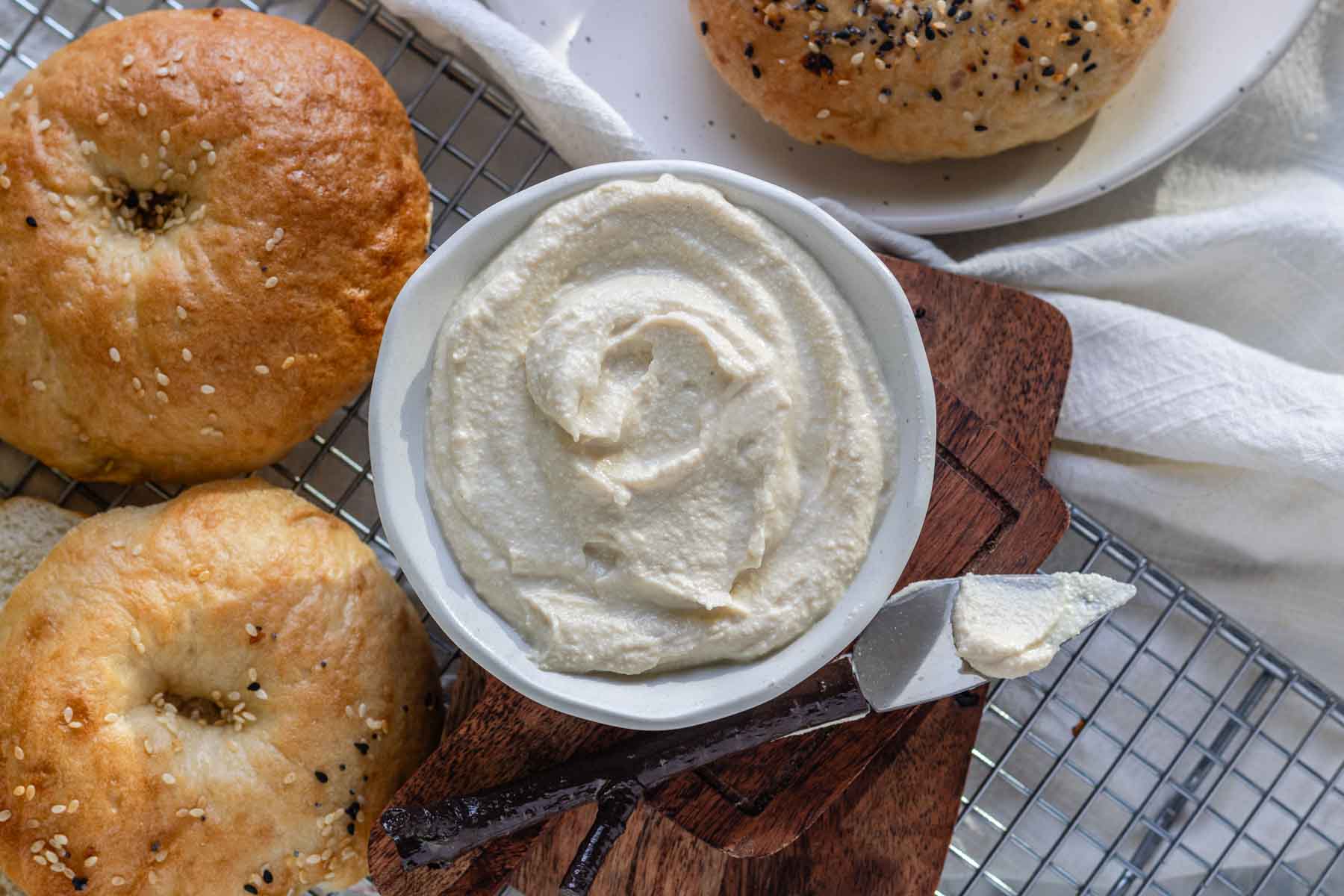 Vegan cream cheese in a bowl on a wooden board beside bagels.