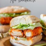 A buffalo tofu sandwich with lettuce on a white plate with pickles, with black text above.