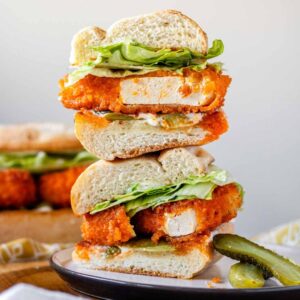A buffalo tofu sandwich with lettuce, stacked on a white plate with pickles.