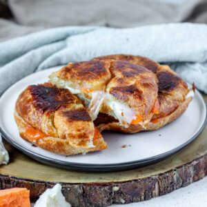A croissant grilled cheese cut in half with another croissant on a white plate on a wooden board.