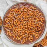 Peanut butter pie in a glass pie dish topped with chocolate ganache and pretzel pieces.