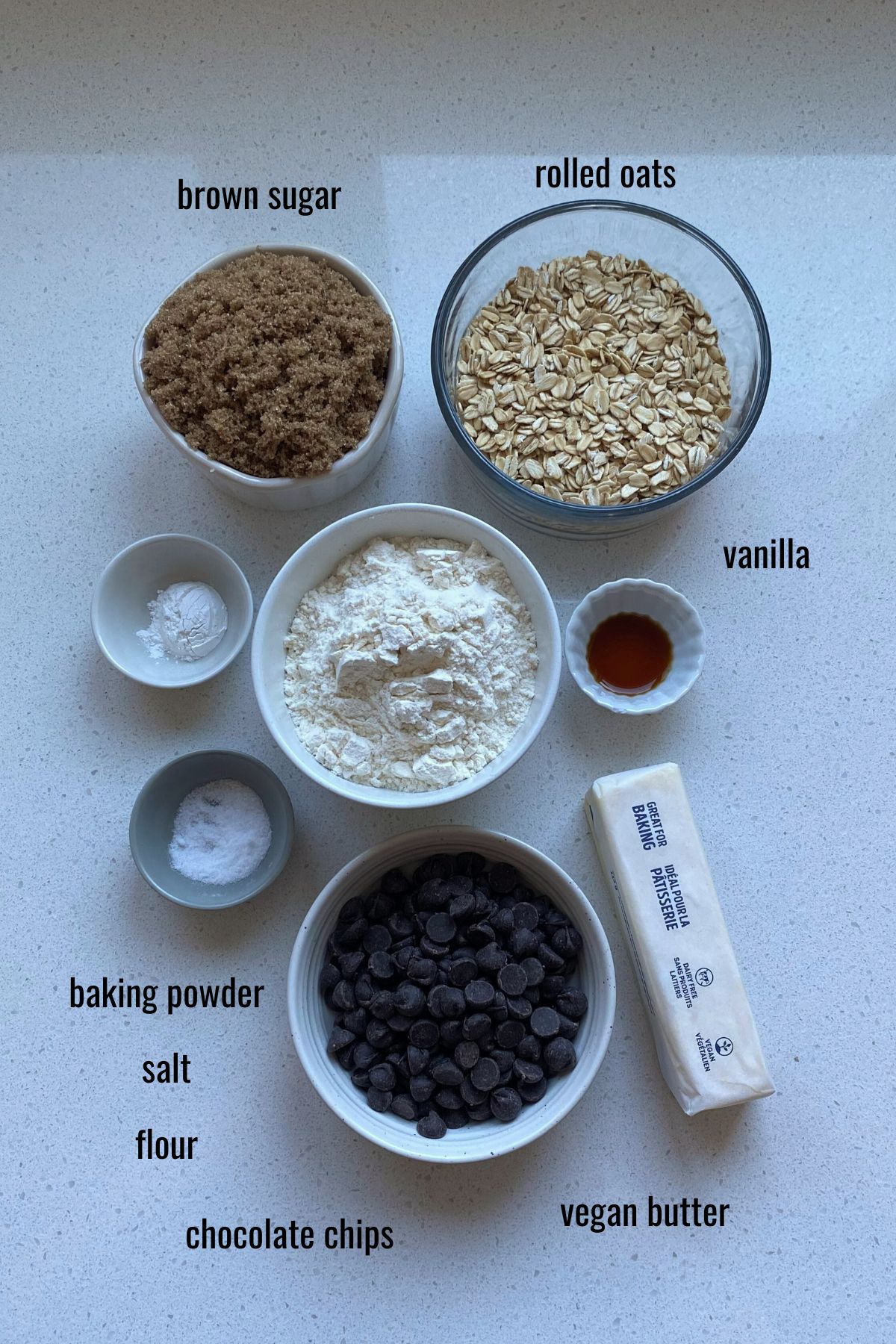 Vegan oatmeal chocolate chip cookies ingredients in dishes on a white counter top.