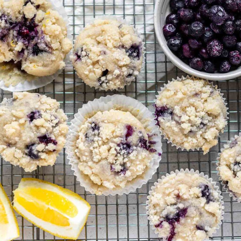 An overhead view of lemon blueberry muffins on a wire rack with lemon wedges and a bowl of blueberries.