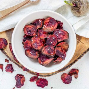 Beet chips in a white bowl on a wooden platter with a white cloth.