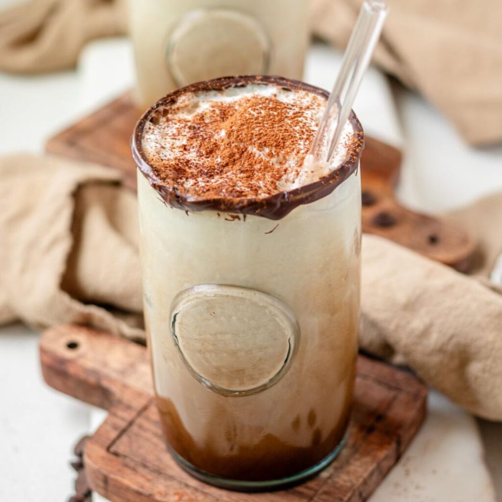 A close up of an iced chocolate latte with a chocolate rip, cocoa powder garnish, and glass straw.
