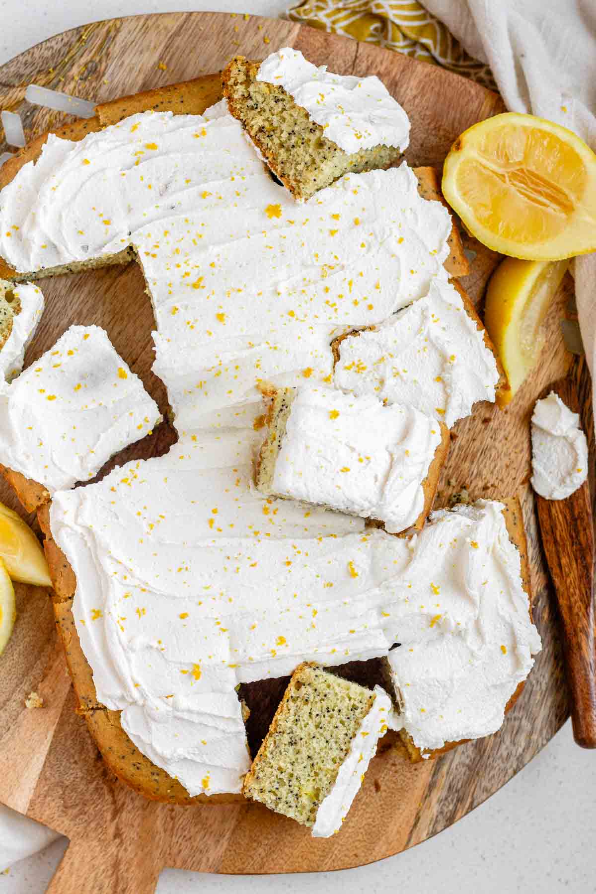 A frosted vegan lemon poppy seed cake sliced into squares on a wooden board.