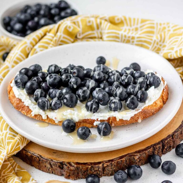 Toast topped with goat cheese, blueberries and honey on a white plate.