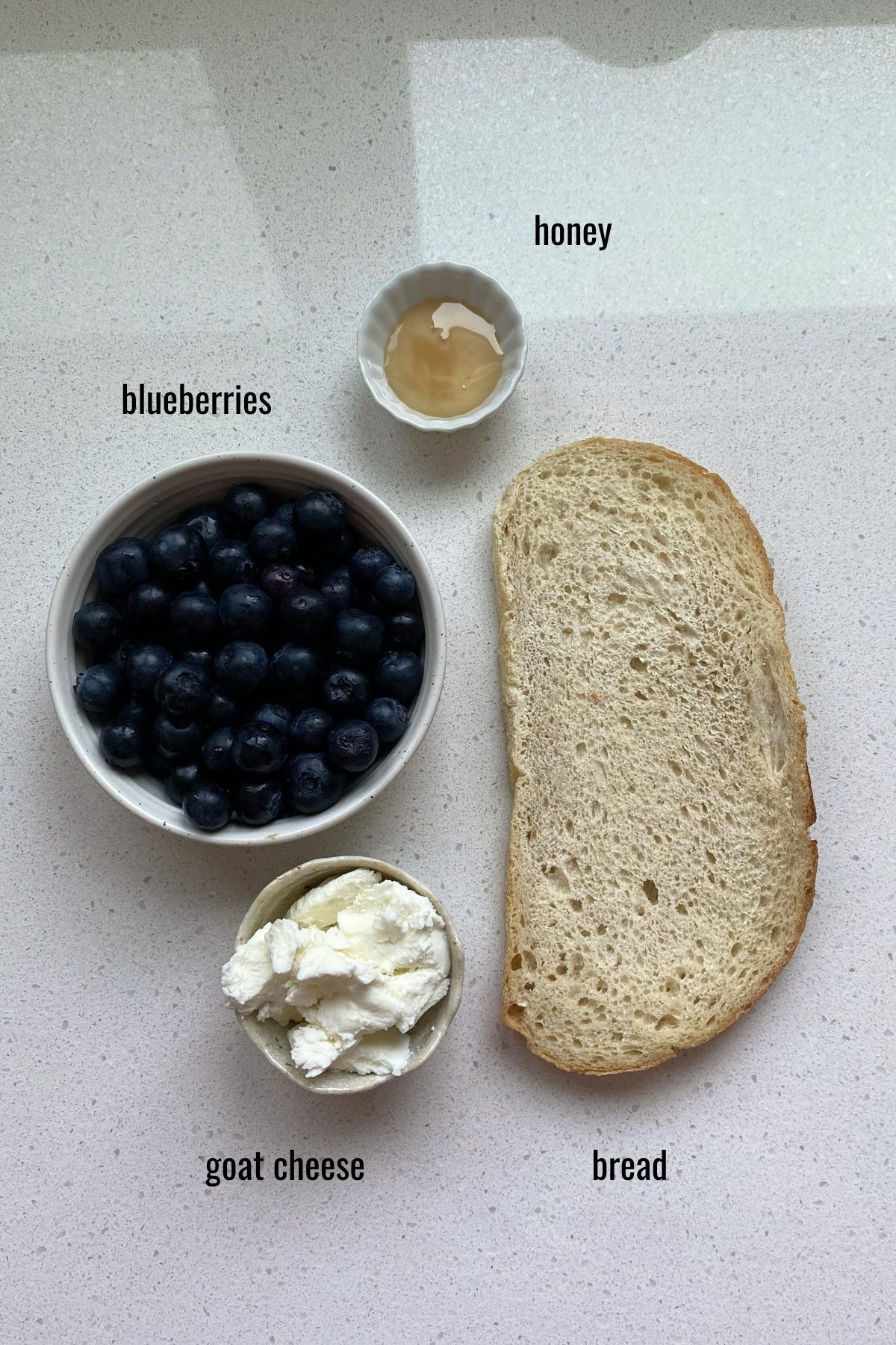Blueberries, honey and goat cheese in bowls with a piece of bread on a white counter.