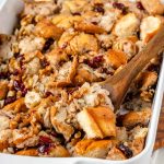 Stuffing with walnuts and cranberries in a white baking dish with a wooden spoon.