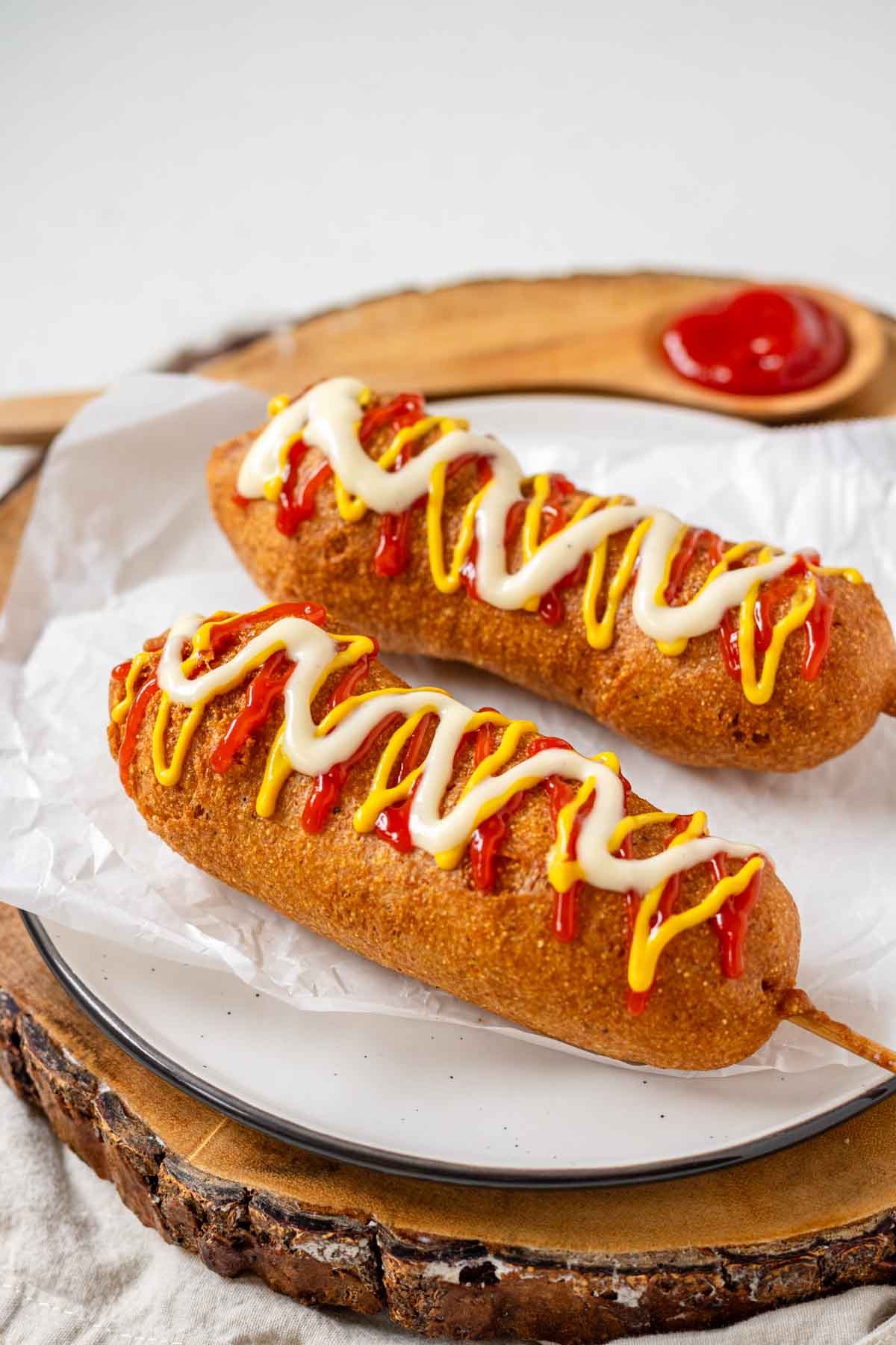 Two vegan corn dogs topped with ketchup, mustard, and vegan mayo on a plate with parchment paper.