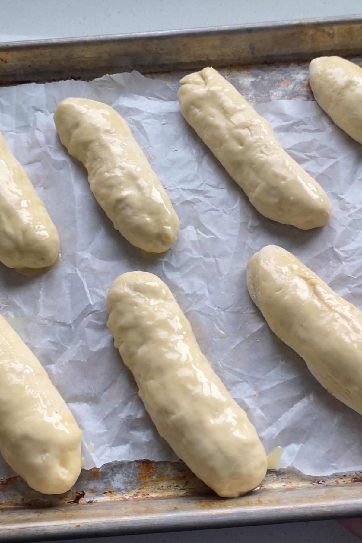 Formed hot dog bun dough brushed with butter on a baking sheet.