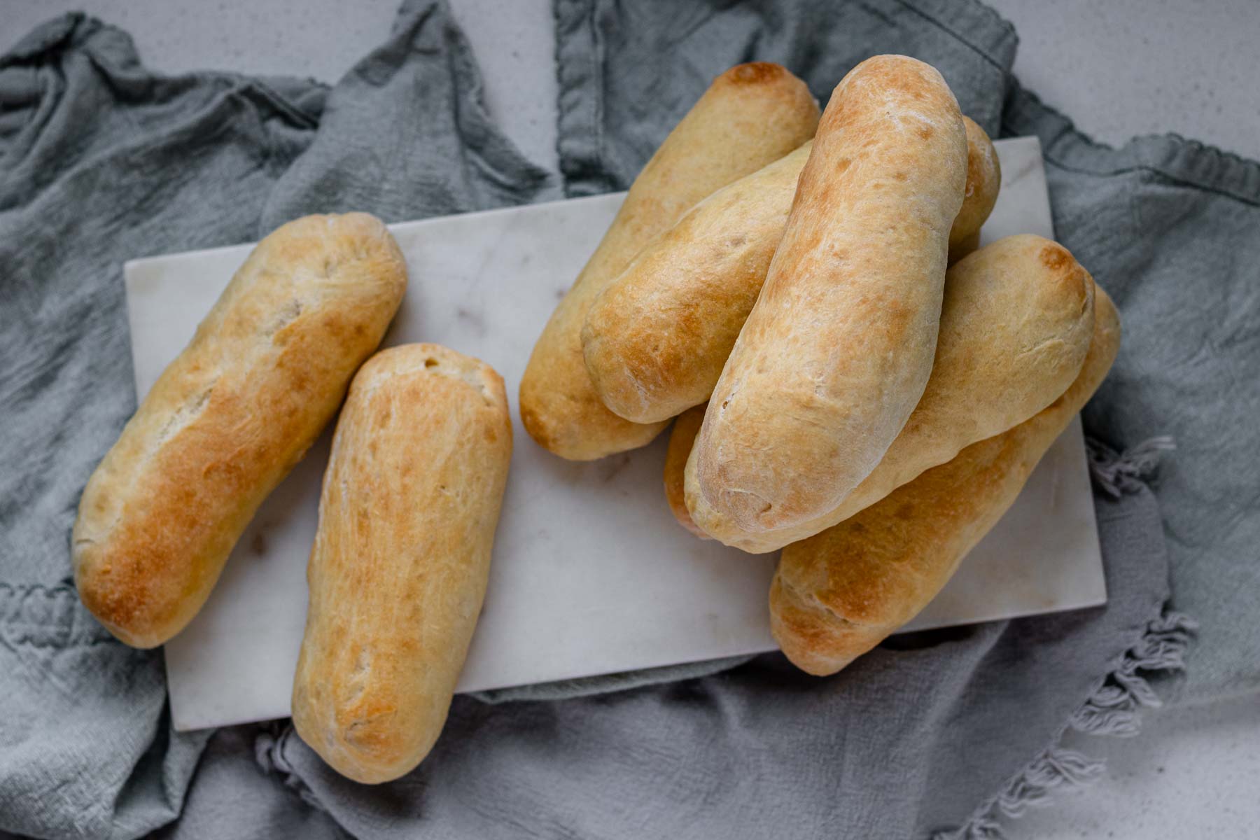 Top down view of hot dog buns stacked on a white marble slab on top of grey cloth napkins.