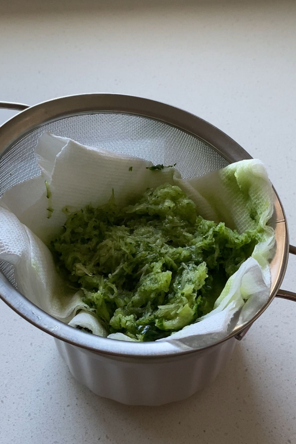 Shredded cucumber in a paper towel lined sieve on top of a small white bowl.
