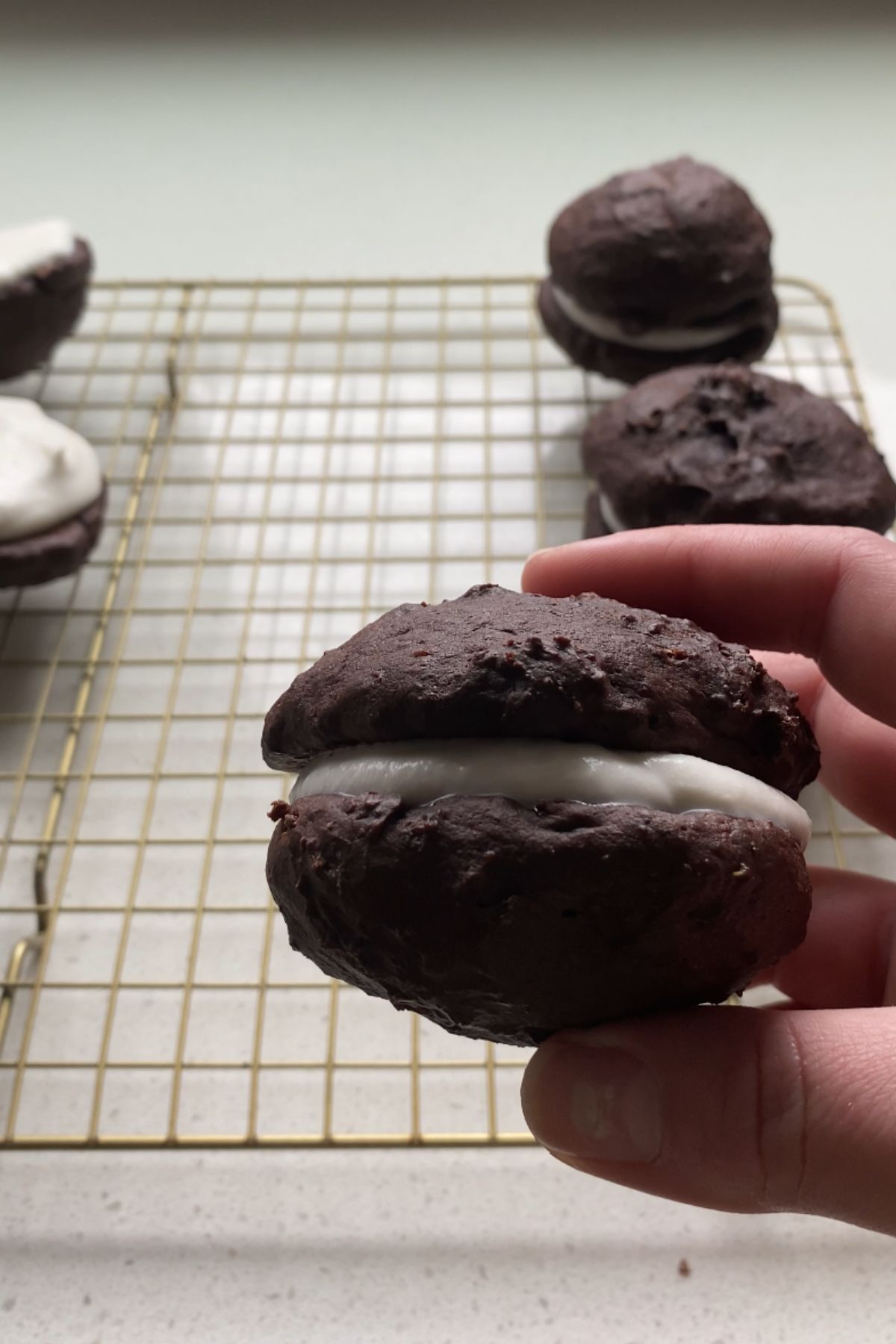 A hand holding a vegan whoopie pie above a cooling rack with more whoopie pies.
