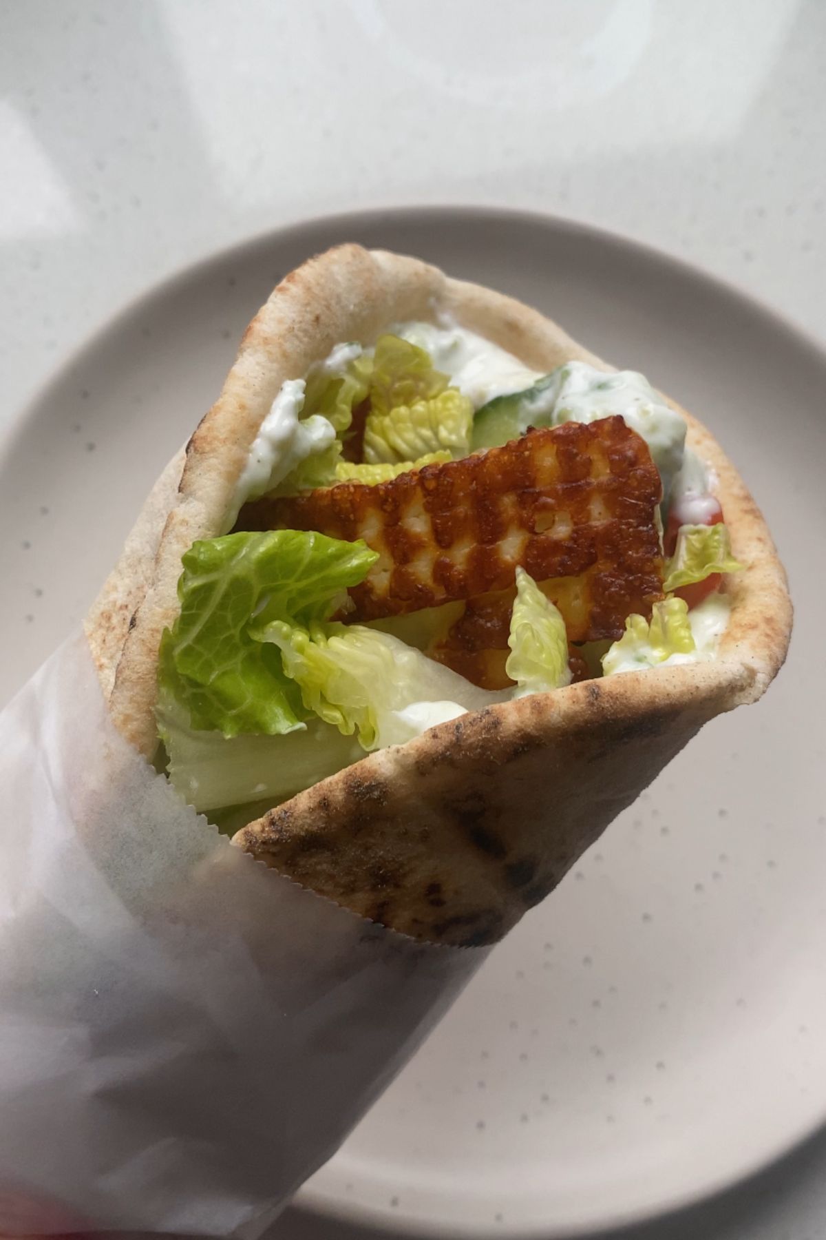 A halloumi gyro wrapped in parchment paper above a white plate.
