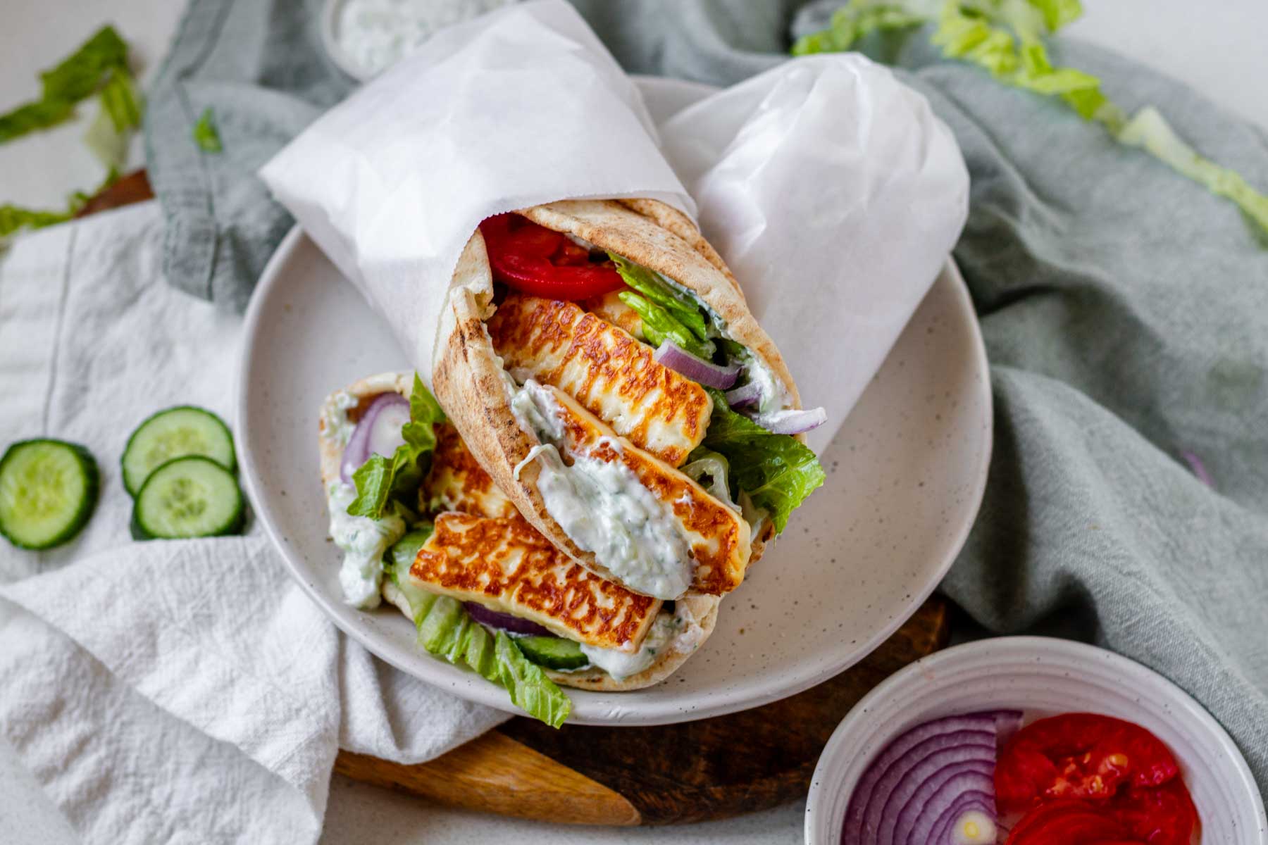 A halloumi gyro wrapped in parchment paper on a white plate surrounded by cloth napkins and veggies.