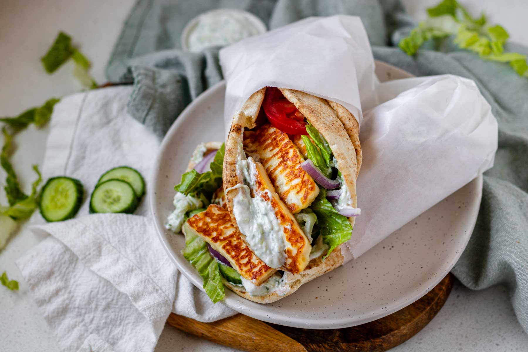 A halloumi gyro wrapped in parchment paper on a white plate surrounded by cloth napkins and veggies.