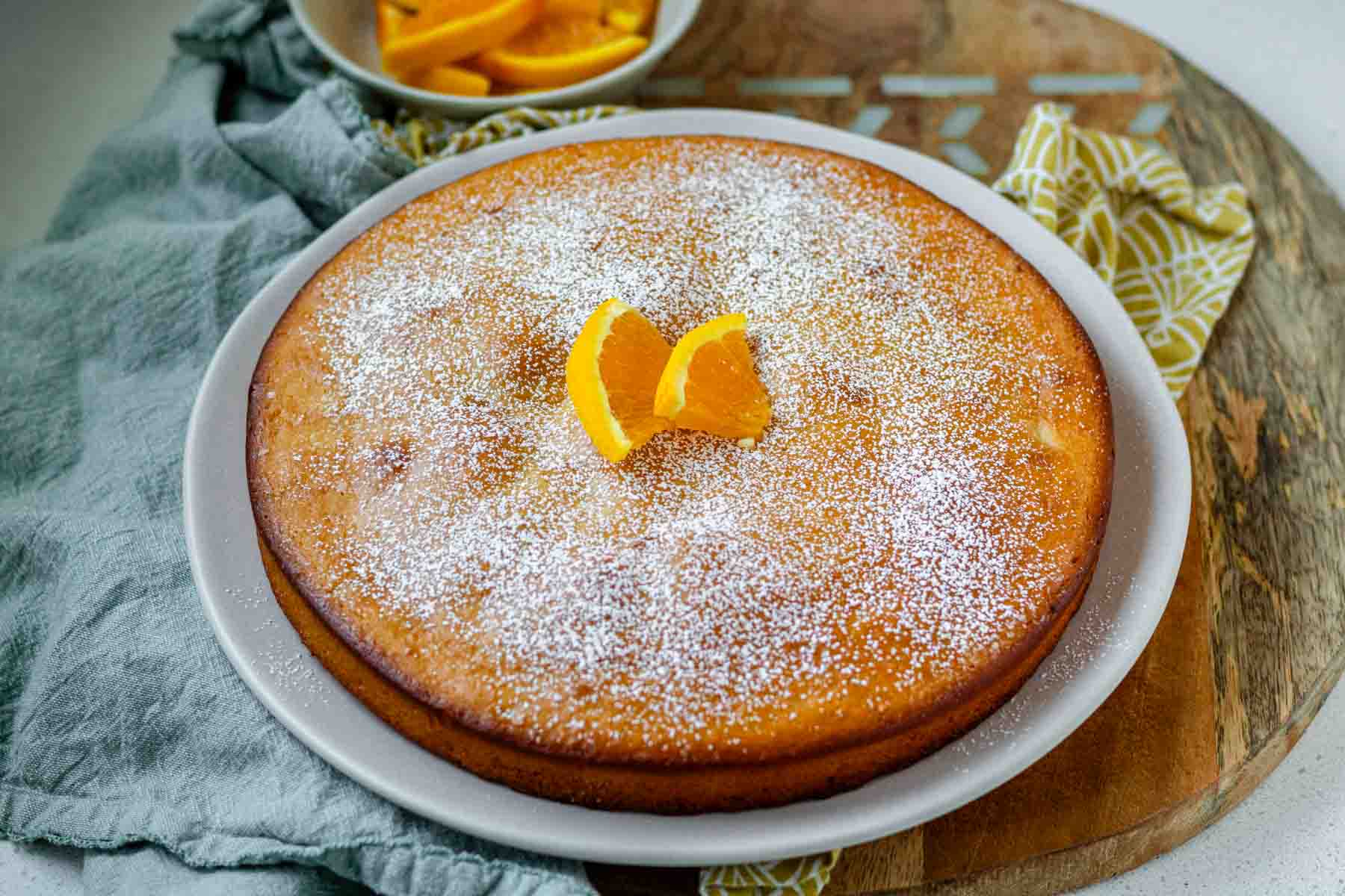An orange blossom cake on a white plate topped with powdered sugar and orange slices.