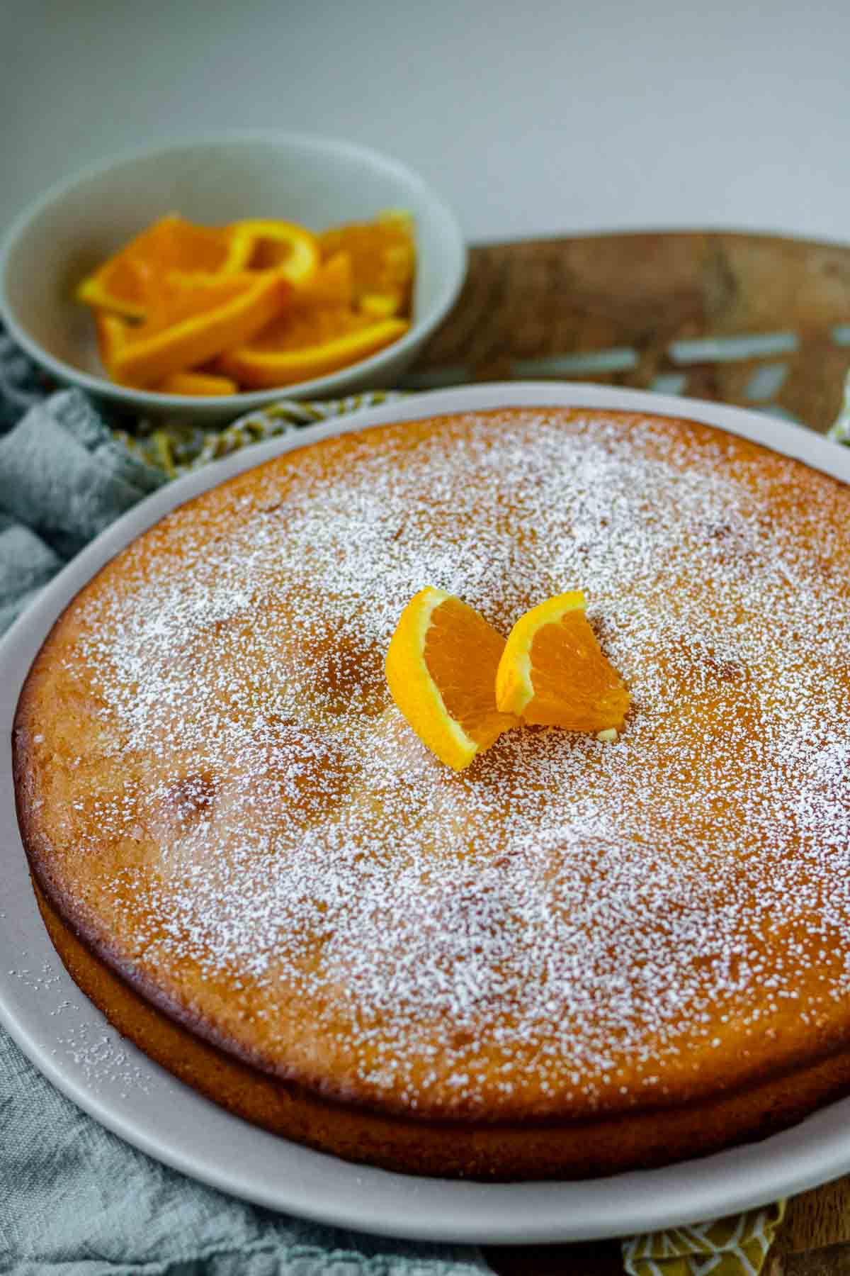 An orange blossom cake on a white plate topped with powdered sugar and orange slices.