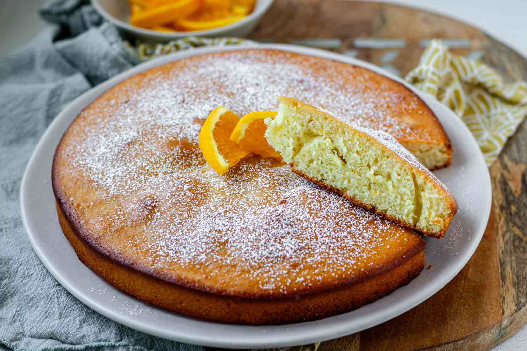An orange blossom cake topped with powdered sugar and orange slices, with a slice resting against the cake.
