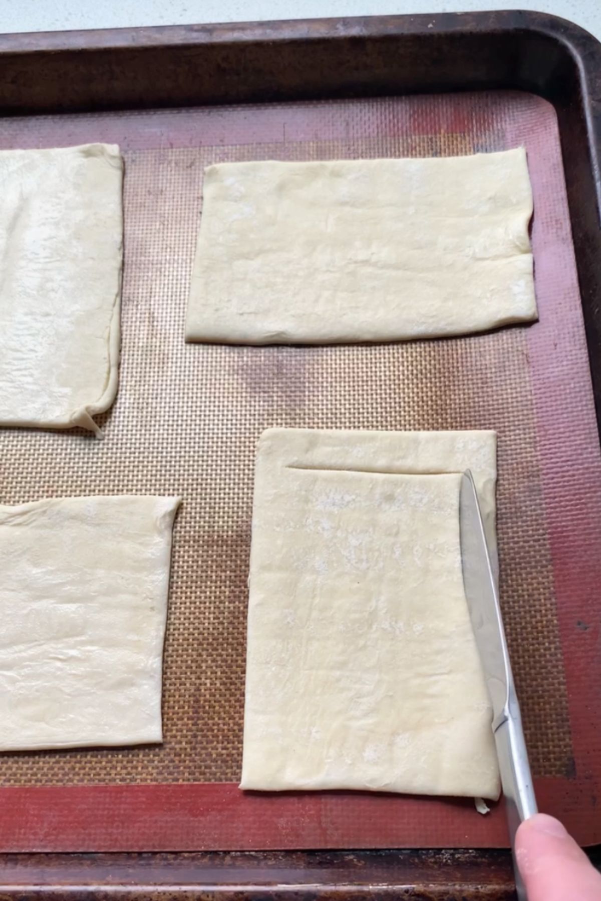A butter knife tracing a border onto puff pastry rectangles on a baking sheet.