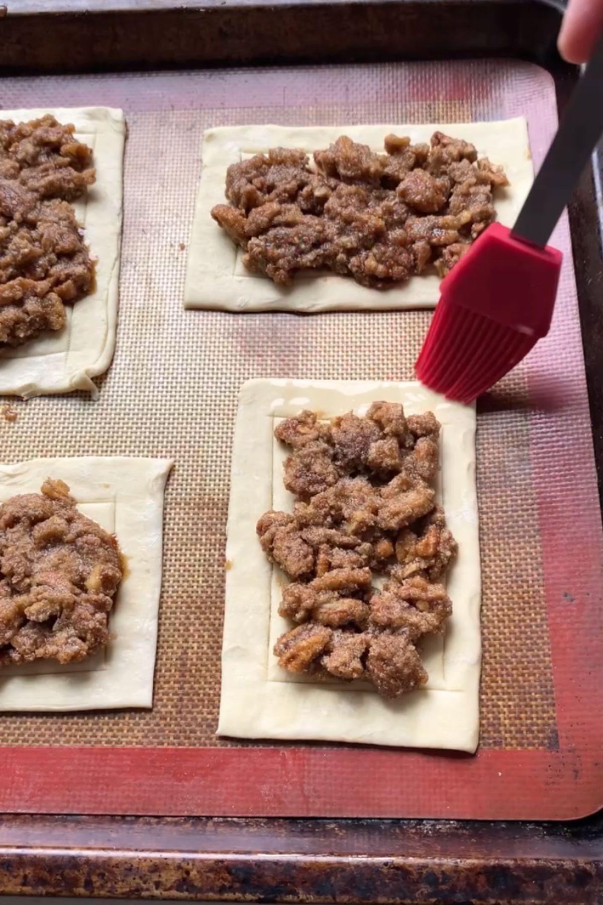 Unbaked maple pecan danishes on a baking sheet with a pastry brush brushing the edges.